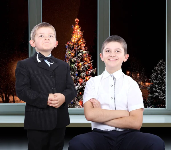 The younger brother in a suit and with a butterfly on her neck, and older brother in a shirt on a background of christmas trees outside the window
