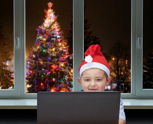 Garçon regarde un ordinateur portable sur fond d'arbre de Noël décoré devant la fenêtre — Photo