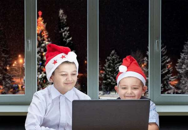 Zwei Brüder sitzen neben dem Laptop auf dem Hintergrund von Weihnachtsbäumen vor dem Fenster — Stockfoto