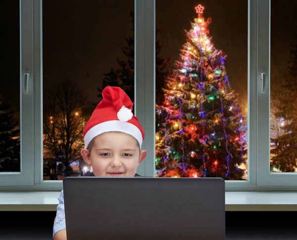 Kleiner Junge sitzt neben einem Laptop in der Mütze des Weihnachtsmannes — Stockfoto