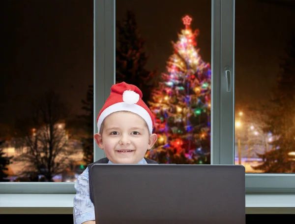 Fröhlicher Junge sitzt neben dem Computer auf dem Hintergrund geschmückter Weihnachtsbaum vor dem Fenster — Stockfoto