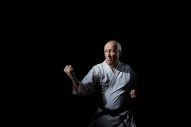 Yaşlı bir erkek sporcu siyah bir arka planda karate duruşunda duruyor.