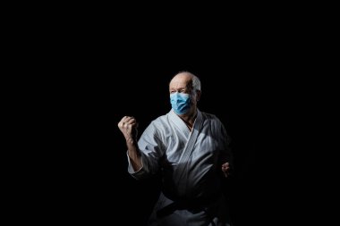 Tıbbi bir maske takan yaşlı bir erkek sporcu, siyah bir arka planda bir blokta antrenman yapıyor.