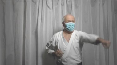 Beyaz karategi ve tıbbi maske takan yetişkin bir sporcu elleriyle yumruk ve blok talimi yapıyor.