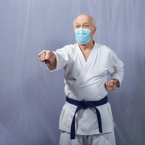 An old athlete with a blue belt and in a medical mask trains a punch arm