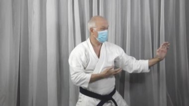 Karategi ve medikal maskeli yaşlı bir erkek sporcu resmi karate egzersizleri yapıyor.