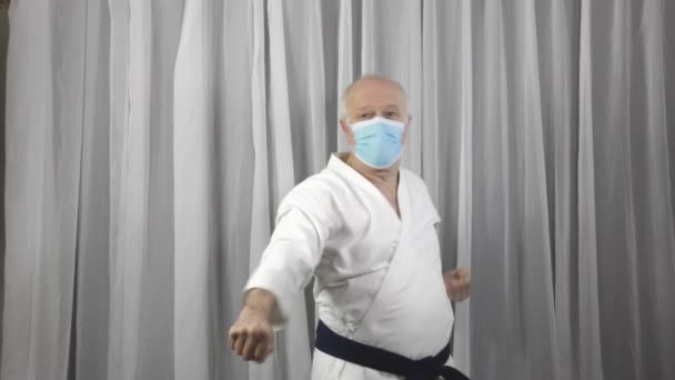 In Karategi und medizinischer Maske macht ein alter männlicher Athlet mit den Händen auf der unteren Ebene Blöcke