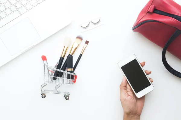 Žena ruka drží mobilní zařízení s nákupní vozík a kosmetické předměty a laptop na bílém pozadí, pohled shora online nákupní koncept — Stock fotografie