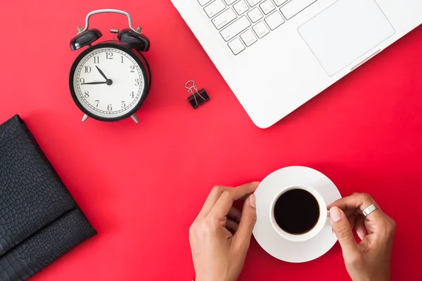 Visão superior do espaço de trabalho, mão da mulher segurando xícara de café, despertador e computador portátil branco no fundo de cor vermelha — Fotografia de Stock