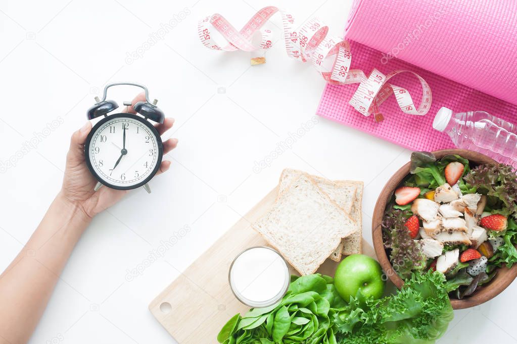 Salad bowl, whole wheat bread, milk, measuring-tape, yoga mat and woman's hand holding alarm clock, Healthy diet concept