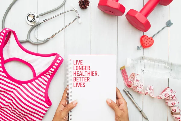 Woman\'s hands holding book, fitness equipments and stethoscope on white wooden background, top view, Healthy lifestyle concept