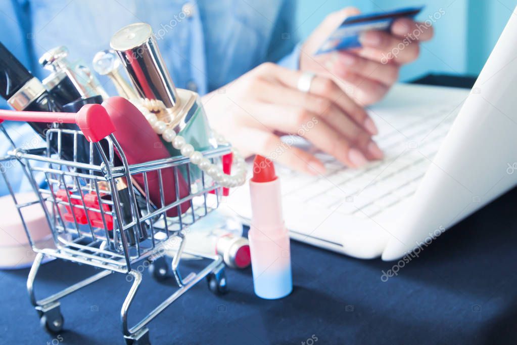 Cosmetics and beauty items in shopping cart with woman using laptop computer for online shopping in background 