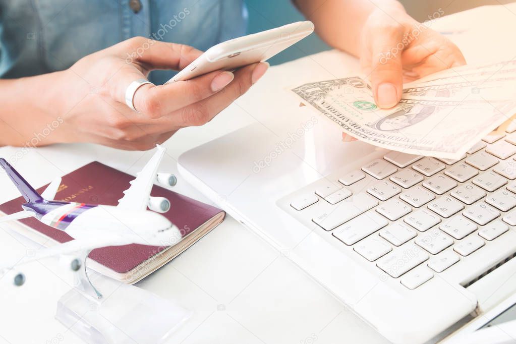 Woman using smartphone and holding dollar bills on other hand, Airplane and passport on table with laptop computer 