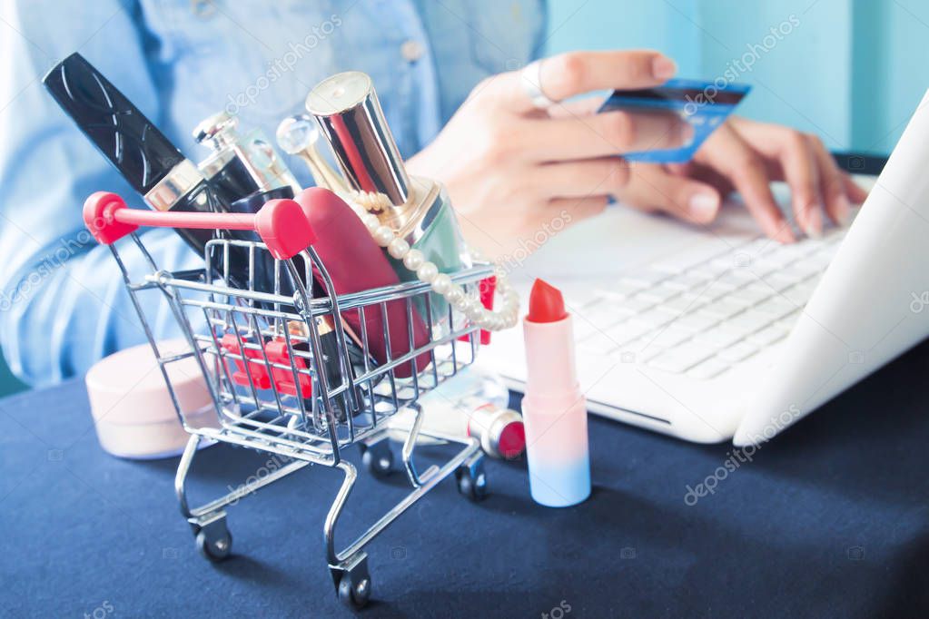 Cosmetics and woman's essentials in shopping cart with woman using laptop and credit card for online shopping 
