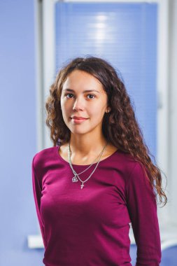 Mor kazak giyen ve kameraya bakan kıvırcık saçlı genç bir kadının portresi.