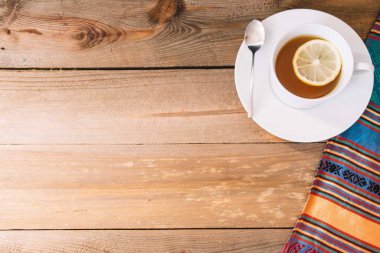 Beyaz fincanda yeşil çay ve beyaz bir seramik tabağın tepesinde bir dilim limon ve ahşap bir arka planı ve renkli bir peçetesi olan bir kaşık var.