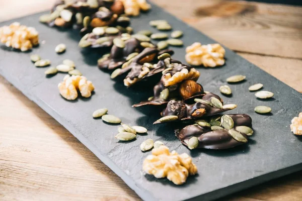 Healthy snack made of almonds, walnuts, hazelnuts and pumpkin seeds covered in dark chocolate standing on a slate dish on a wooden base and black background — Stock Photo, Image