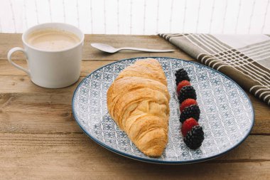 Akdeniz desenli bir tabakta servis edilen kruvasanın yanında böğürtlen ve ahududu ile birlikte bir kahve, kaşık ve ahşap bir tabanda kahverengi ve beyaz bir peçete servis edilir.