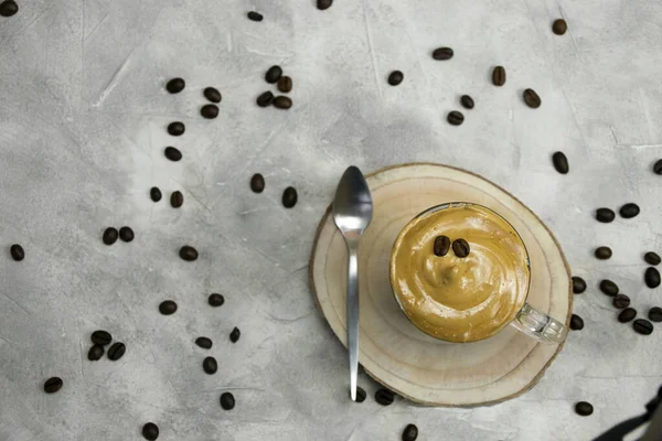 Top view of whipped instant coffee con a crystal mug with a spoon on a wooden coaster on a grey background with coffee beans sprinkled and blurred background. Dalgona coffee concept