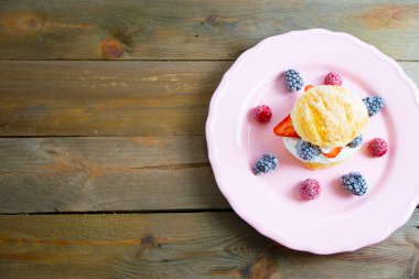 Krem şanti, ahududu, böğürtlen ve böğürtlen ile dolu soluk pembe bir tabakta ahşap bir arka planda poğaça. Çöl konsepti.