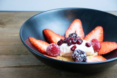 Dilimlenmiş çilek, kurabiye kırıntısı, krema, nar tohumu, böğürtlen ve çilek dolu siyah mat tabağı..