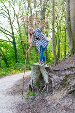 Genç bir kadın ormanda büyük bir ağaç kütüğüne gülerek ayakta duruyor ve bir bastona yaslanıyor. Konum: Almanya, Kuzey Ren Vestfalyası