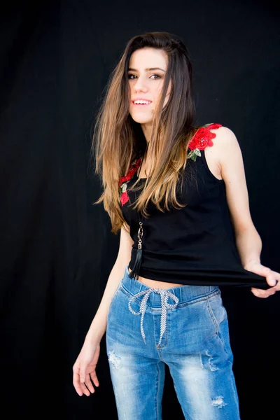 Νεαρή κοπέλα με αμάνικο t-shirt με λουλουδάτο μοτίβο για τον ώμο και μοντέρνα τζιν, με μακριά καστανά μαλλιά. Μαύρο φόντο — Φωτογραφία Αρχείου