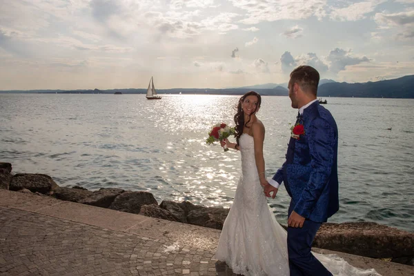 若い夫婦 トーリ ベナコの湖畔に沿って散歩 イタリア 花嫁はレースドレスを着て 新郎はエレガントな青いドレスを着ています 湖の背景に雲や水面への反射 — ストック写真