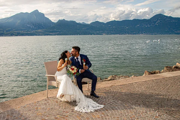 Young married couple, sitting on a bench with ice cream in hand along the lake shore in Torri del Benaco, Italy. Lake background with clouds and reflections on the water.