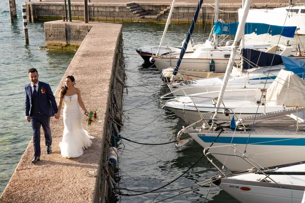 Young married couple strolling at the dock of the lake in Torri del Benaco, Italy. The bride is wearing a lace dress, the groom is wearing an elegant blue dress.