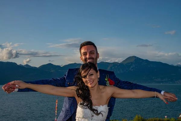 Young married couple with arms raised, happy smiling. Lake Garda, Torri del Benaco, Italy. Background with mountains and white clouds.