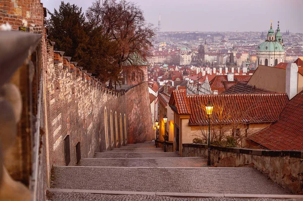 Old Castle Steps (stairs) that lead out of Lesser Quarter Street up to the eastern gate of Prague Castle captured in sunset light with pink sky and the panoramic view to the city.