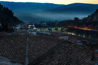 Evening Berat, town is the south of Albania, with its hills, covered with mist, illuminated Gorica Bridge over Osum river and old buildings with tile roofs. clipart