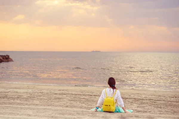 Młoda kobieta siedzi na piaszczystej plaży patrząc na morzu i zachód słońca — Zdjęcie stockowe