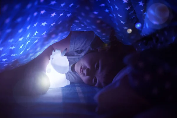 Menino abraçando uma lâmpada de unicórnio de brinquedo sob um cobertor com estrelas — Fotografia de Stock