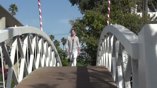 リゾートタウンの木製の橋を歩いてスタイリッシュなハンサムな若い男 — ストック動画