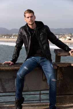 Çekici genç adam bir tatil köyünde okyanusun üzerinde bir iskelede oturuyor, seyahat ve turizm konsepti.