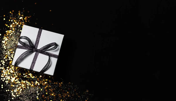 White gift box with silver ribbon and gold confetti and sparkles on black background. Top view, copy space. ストック画像