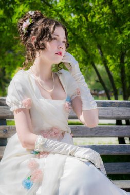 Portrait of a young bride woman in a historical white dress with a book in hands outdoors in a park on a bench. clipart
