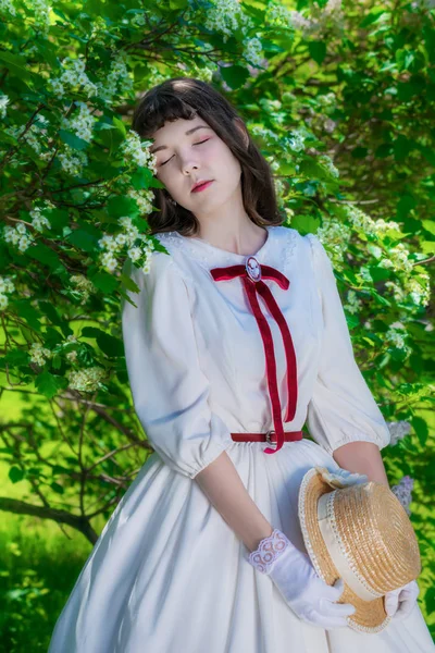 Niña en un vestido blanco y sombrero de paja cerca de flores de espino — Foto de Stock