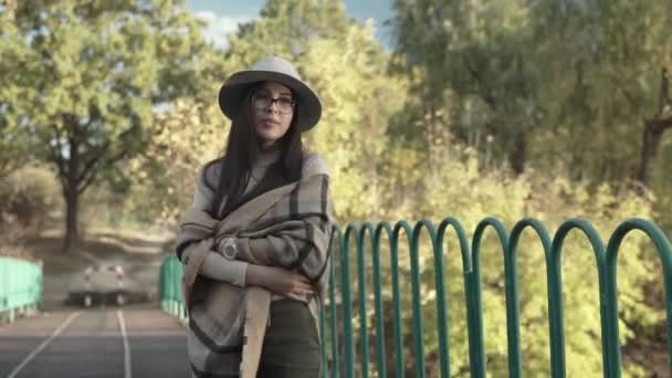 一个戴着帽子的迷人的年轻女子在秋天的公园里沿着湖上的一座桥散步 美丽的女子在公园散步时在黄叶上的秋天肖像 — 图库视频影像