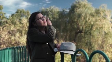 Şapkalı çekici genç bir kadın bir sonbahar parkında bir gölün üzerindeki köprü boyunca yürüyor. Sonbaharda parkta yürürken sarı yaprakların üzerinde güzel bir kadının sonbahar portresi.
