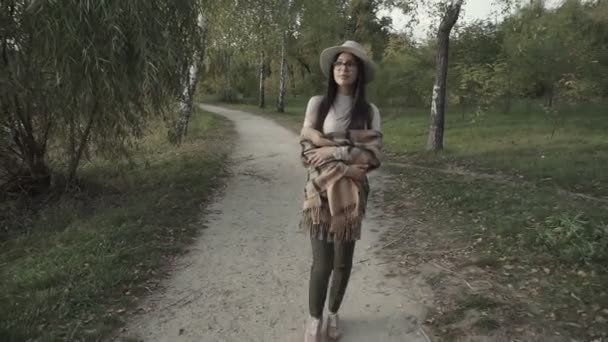 一个戴着帽子和眼镜的年轻的黑头发姑娘在秋天的公园里散步 — 图库视频影像