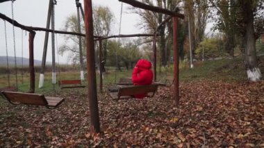 Sonbahar parkında kırmızı ceketli bir kadın salıncakta sallanıyor.