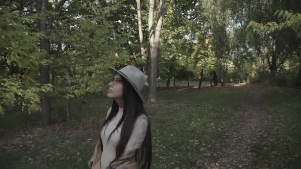 一个戴着帽子和眼镜的年轻的黑头发姑娘在秋天的公园里散步 — 图库视频影像