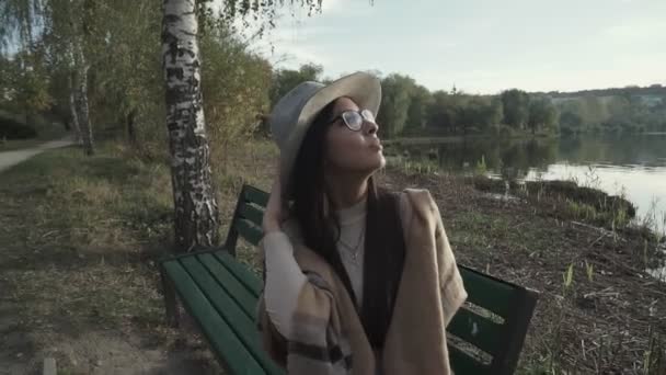 放松点 待在这里 年轻的女人 戴着帽子坐在长椅上 望着远方的落日和湖面 — 图库视频影像