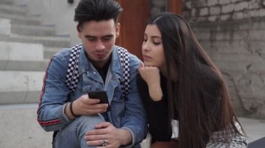 Genç bir çiftin portresi parkta bir bankta oturur ve akıllı telefondaki fotoğraflara bakar.