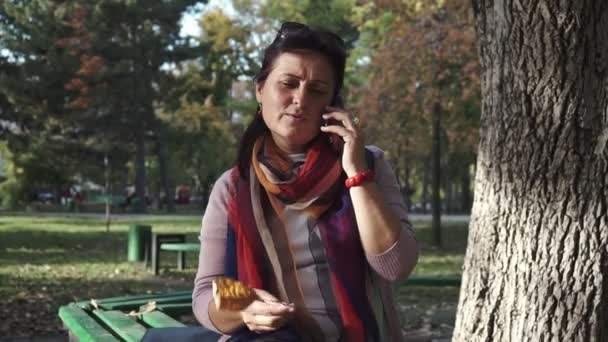 坐在公园的长椅上 笑着用智能手机说话的黑发女人 — 图库视频影像