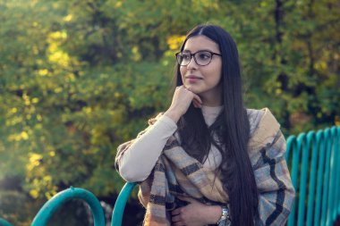 Uzun saçlı ve gözlüklü, duygusal olarak mutlu esmer kadına yakın durun. Kuaförden sonra mükemmel makyajlı, parkta poz veren güzel bir model. Moda ve güzellik kavramı.