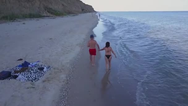 サンセットのビーチを歩く男と女ビーチで楽しい休暇を過ごしているカップルの空中撮影を追跡します ビーチで夕日の空のビーチを歩くカップル — ストック動画